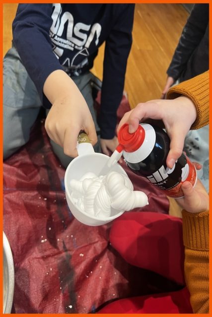 A child spraying shaving cream into a measuring cup that another child is holding